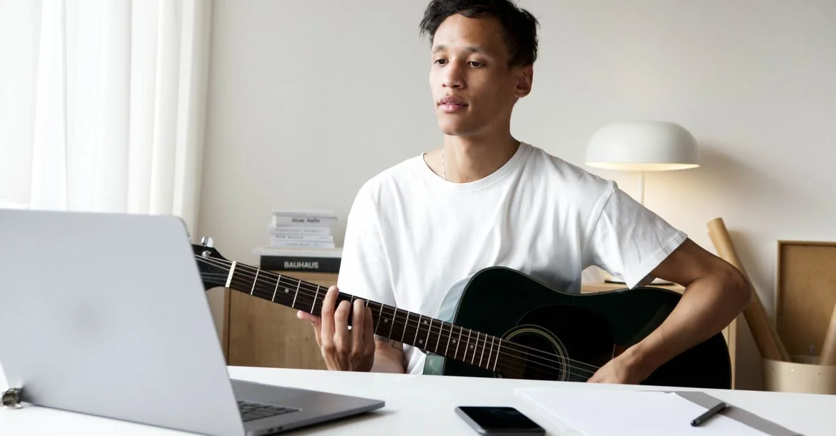 Homme jouant de la guitare acoustique tout en utilisant un ordinateur portable dans une pièce bien éclairée, idéal pour des concepts de créativité musicale.